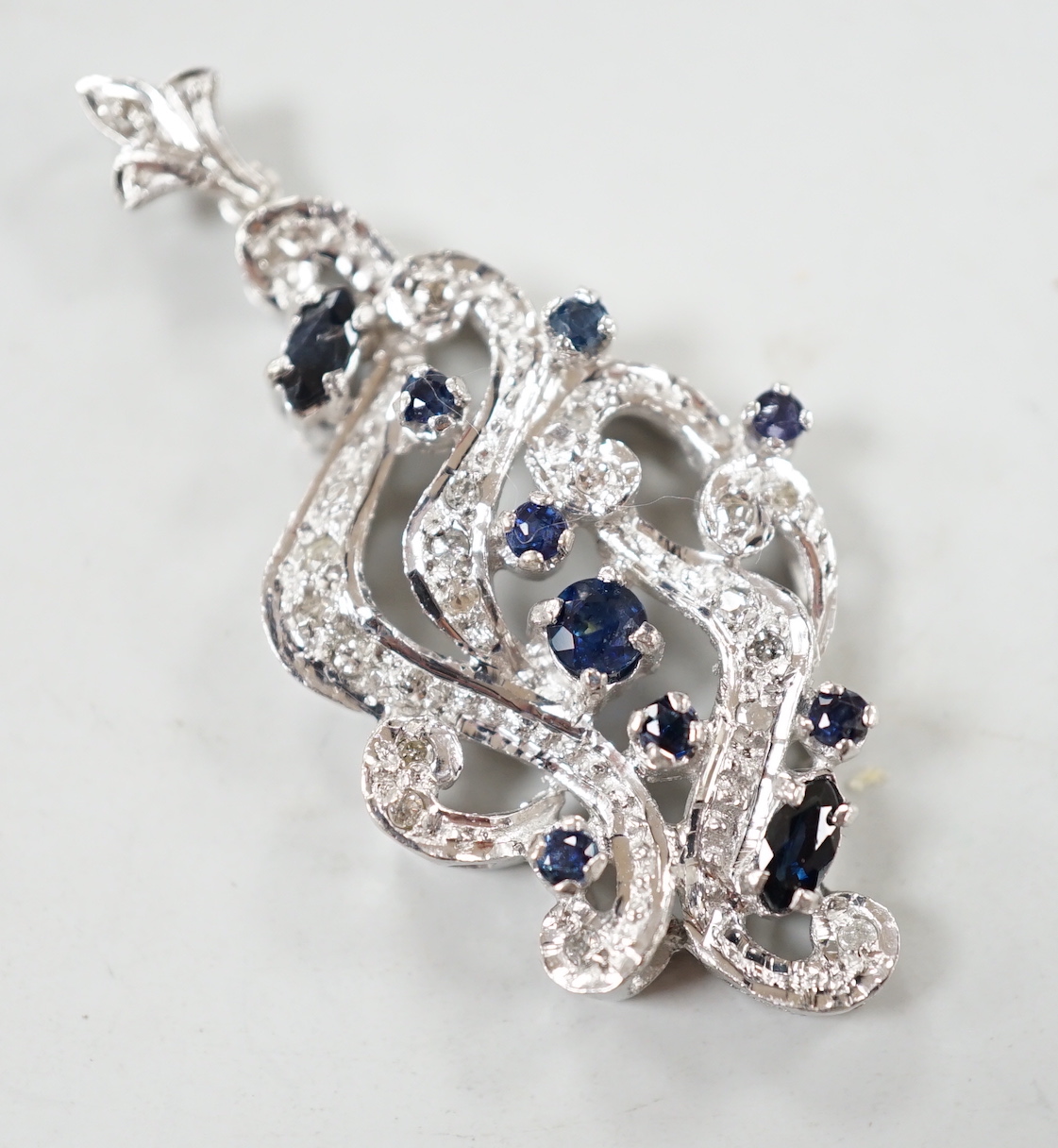 A modern 18k white metal sapphire and diamond chip cluster set scroll pendant, overall 47mm, gross weight 7.7 grams.